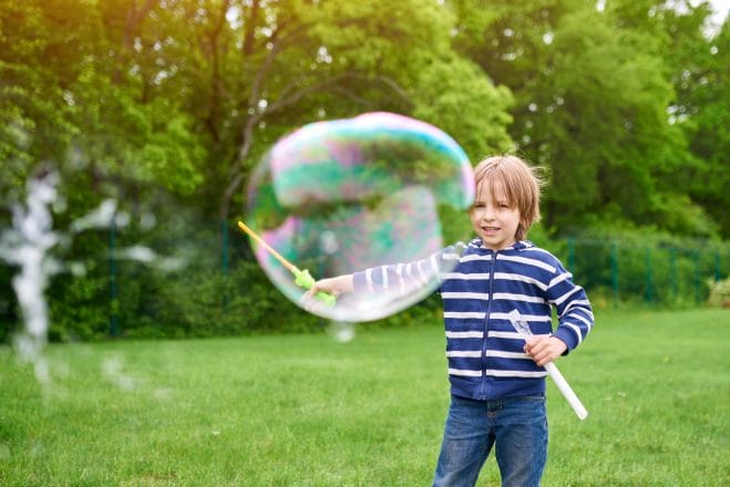 Outdoors portrait of cute preschool boy blowing soap bubbles on a green lawn at the playground