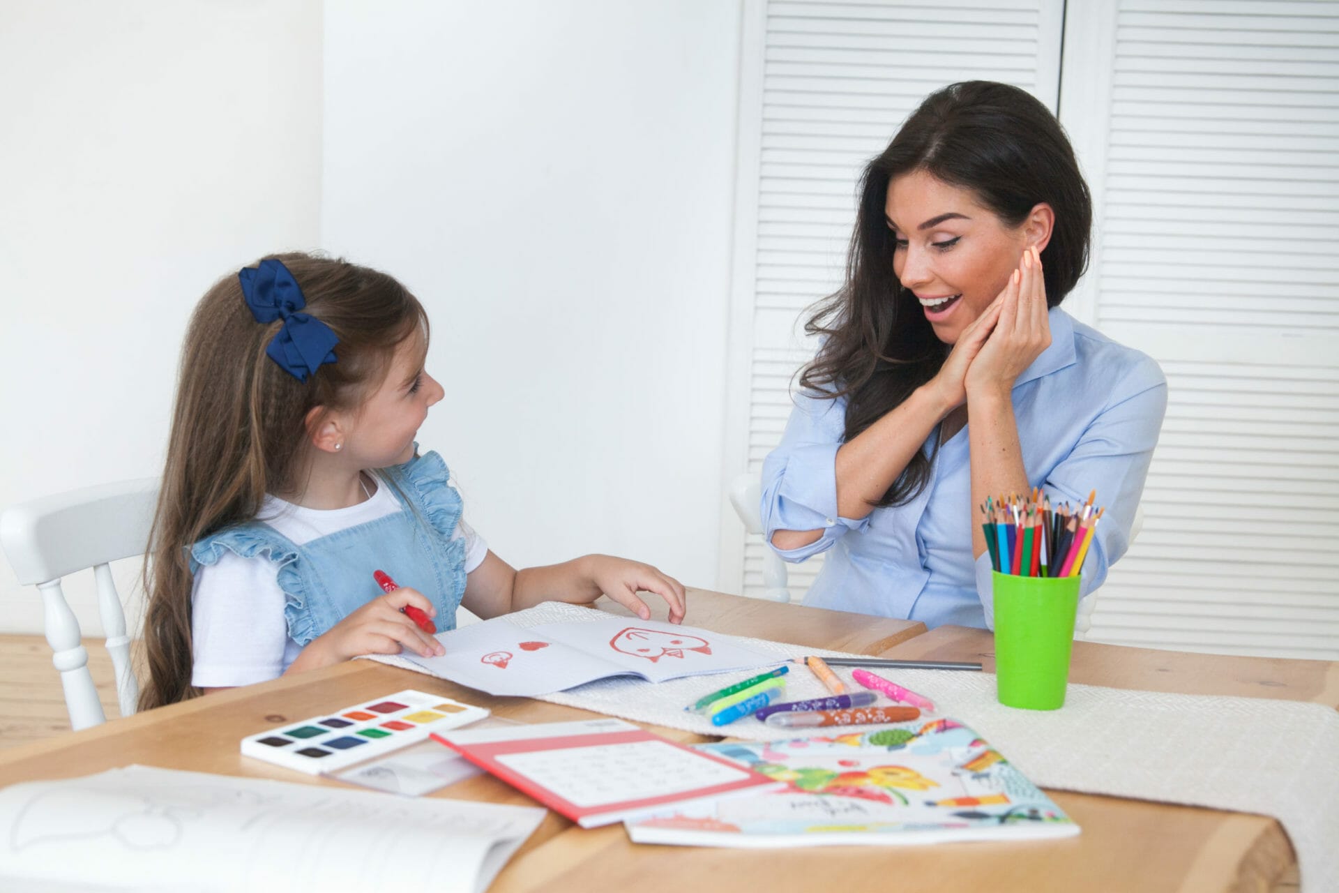 Smiling mother and daughter preparing for lessons and draws at the table with pencils and paints